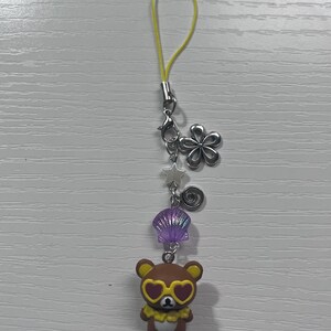 Puede incluir: Un amuleto de oso marr&oacute;n con gafas de sol en forma de coraz&oacute;n y un collar de flores amarillas. El amuleto est&aacute; unido a una cuerda amarilla con un cierre plateado y varios otros amuletos, incluyendo una concha, una estrella y un remolino.