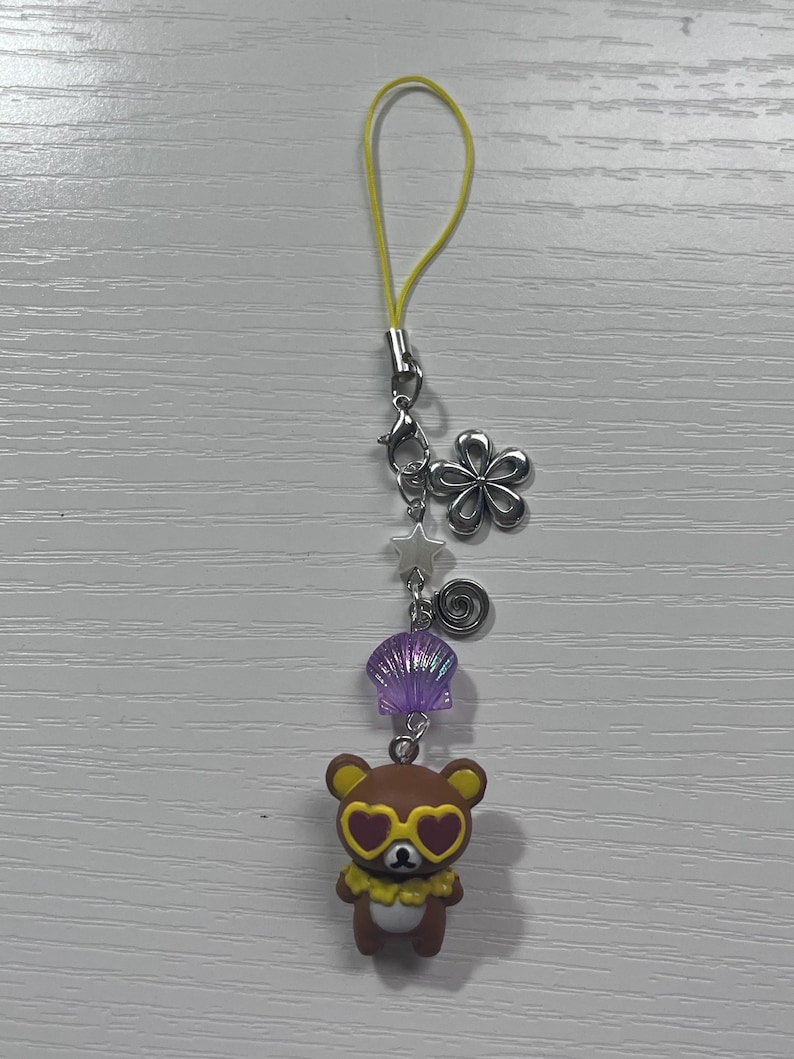 Puede incluir: Un amuleto de oso marr&oacute;n con gafas de sol en forma de coraz&oacute;n y un collar de flores amarillas. El amuleto est&aacute; unido a una cuerda amarilla con un cierre plateado y varios otros amuletos, incluyendo una concha, una estrella y un remolino.