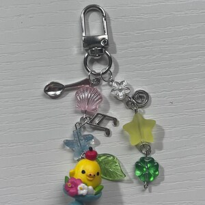 Puede incluir: Un amuleto de pollito amarillo con una cereza roja en la parte superior, sentado en un cuenco azul con flores rosas. El amuleto est&aacute; unido a un llavero plateado con varios amuletos, incluyendo una concha, una cuchara, una estrella, un tr&eacute;bol, una flor y notas musicales.