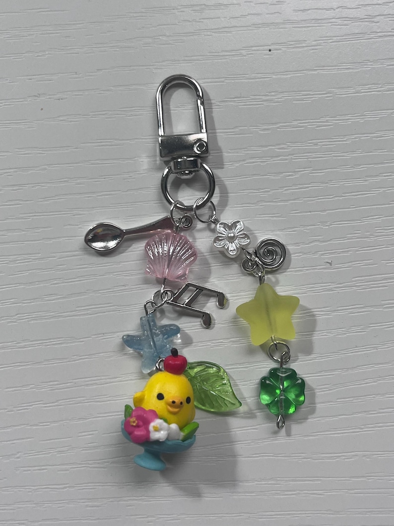 Puede incluir: Un amuleto de pollito amarillo con una cereza roja en la parte superior, sentado en un cuenco azul con flores rosas. El amuleto est&aacute; unido a un llavero plateado con varios amuletos, incluyendo una concha, una cuchara, una estrella, un tr&eacute;bol, una flor y notas musicales.