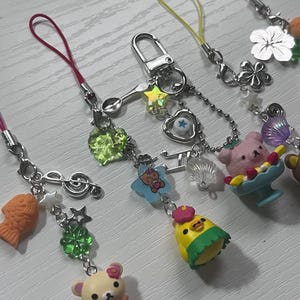 Puede incluir: Una colecci&oacute;n de abalorios para tel&eacute;fono de colores con personajes de dibujos animados, una cuchara, una nota musical, una estrella, un tr&eacute;bol, una concha, un pez y un coraz&oacute;n. Los abalorios est&aacute;n unidos a cuerdas de colores.