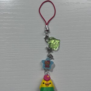 Puede incluir: Un amuleto de pollito amarillo con una flor rosa en la cabeza y una falda de hierba verde. El amuleto est&aacute; unido a una cuerda roja con un cierre plateado y un amuleto de hoja verde. Un amuleto de estrella azul con Hello Kitty tambi&eacute;n est&aacute; unido a la cuerda.