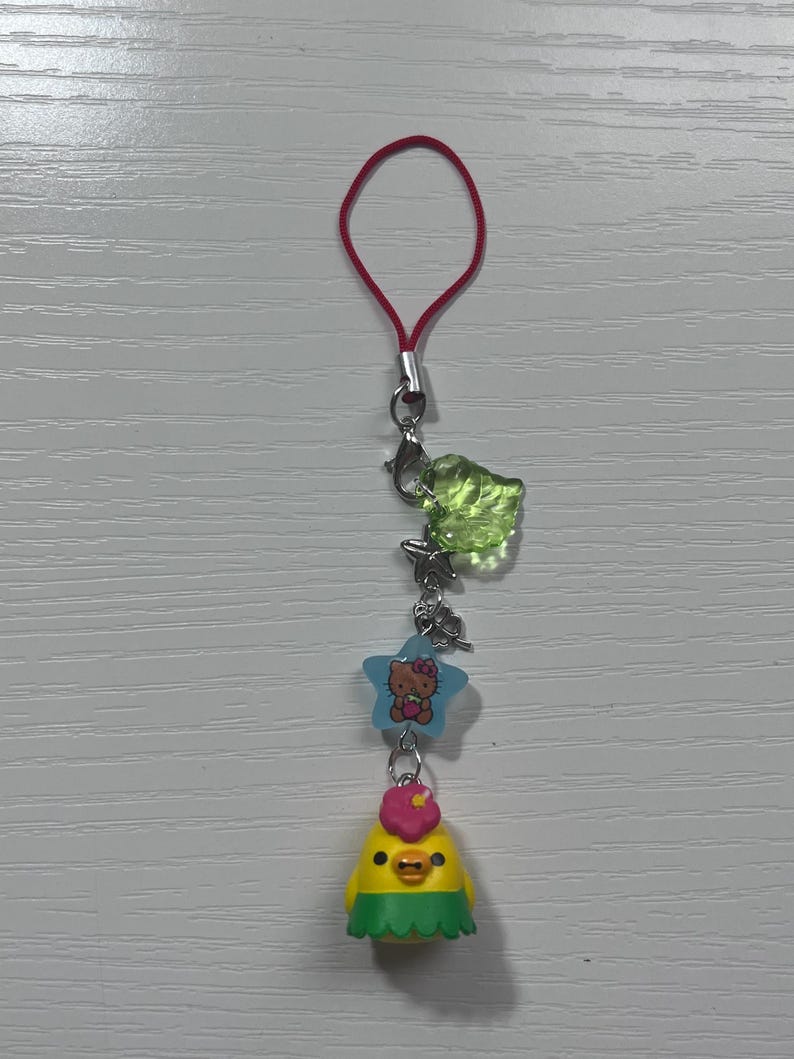 Puede incluir: Un amuleto de pollito amarillo con una flor rosa en la cabeza y una falda de hierba verde. El amuleto est&aacute; unido a una cuerda roja con un cierre plateado y un amuleto de hoja verde. Un amuleto de estrella azul con Hello Kitty tambi&eacute;n est&aacute; unido a la cuerda.