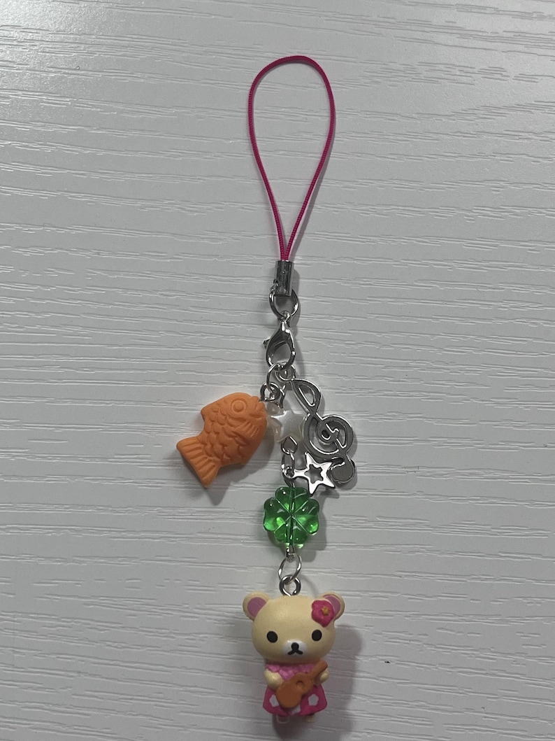Puede incluir: Un llavero de tel&eacute;fono rosa con un dije de oso rosa y blanco con un ukelele, un dije de tr&eacute;bol de cuatro hojas verde, un dije de nota musical plateado, un dije de estrella plateada y un dije de pez naranja.