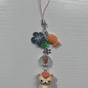 Puede incluir: Un oso marr&oacute;n que lleva una falda de hula verde y una flor en el pelo. El oso est&aacute; unido a un charm de tel&eacute;fono con una cuerda rosa, un charm de Hello Kitty, una estrella verde, una flor azul y un charm de pez naranja.