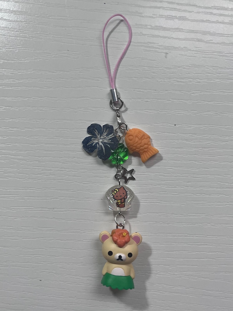 Puede incluir: Un oso marr&oacute;n que lleva una falda de hula verde y una flor en el pelo. El oso est&aacute; unido a un charm de tel&eacute;fono con una cuerda rosa, un charm de Hello Kitty, una estrella verde, una flor azul y un charm de pez naranja.