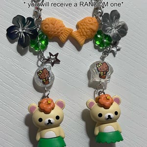 Puede incluir: Dos llaveros con un oso de dibujos animados que lleva una falda de hula verde y una flor en la cabeza. Cada llavero tiene un dije de cuentas de concha diferente, un dije de pez, un dije de flor, un dije de tr&eacute;bol y un dije de estrella. El texto "both keychains have different shell beads * you will receive a RANDOM one*" es visible en la imagen.