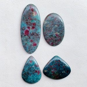 Può includere: Quattro pietre preziose lucide di varie forme: un ovale, una goccia, un triangolo e un ovale arrotondato. Le pietre presentano un colore di base turchese con inclusioni rosse e grigie, creando un effetto marmorizzato. Le pietre sono su una superficie bianca.
