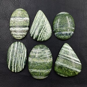 Può includere: Sei cabochon levigati in pietra con motivi verdi e bianchi disposti su due file. Le pietre presentano forme varie, tra cui ovali e a goccia, mostrando distinte bande minerali orizzontali e verticali in tonalità di verde bosco.