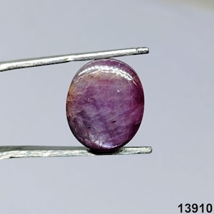 May include: A polished, oval-shaped gemstone held by tweezers. The gem is a deep purple color with lighter streaks and a glossy surface. The gem is likely a ruby or similar precious stone. The number "13910" is visible in the bottom right corner.
