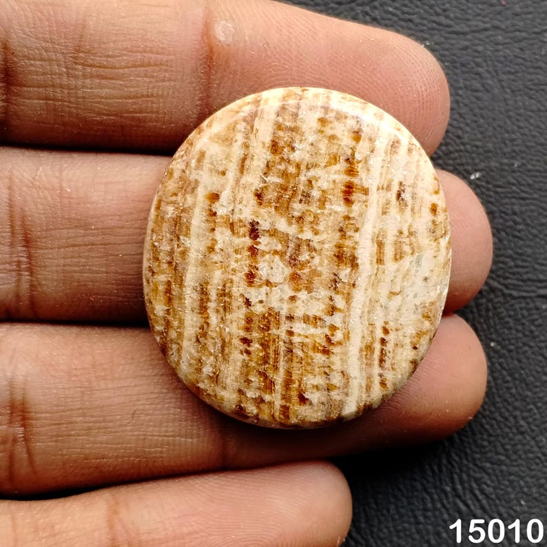 Puede incluir: Una piedra ovalada de color marr&oacute;n claro con un patr&oacute;n a rayas en tonos beige y marr&oacute;n. La piedra pulida tiene un aspecto natural y terroso. La piedra se sostiene en una mano.