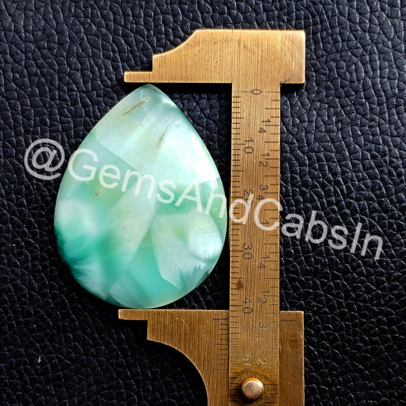 May include: A teardrop-shaped green and white agate cabochon measuring approximately 3.5 cm in length. The cabochon is being measured with a ruler.