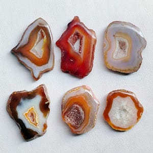 May include: Six polished agate slices in shades of red, orange, brown, and white. Each slice has a unique shape and a crystalline center. The stones are displayed on a white surface, showcasing their natural beauty and intricate patterns.