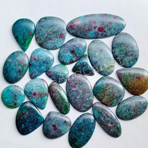 May include: A selection of polished gemstones in diverse shapes, such as oval, teardrop, and irregular forms. The stones display a blend of teal, green, and red tones, with a mottled texture. The gemstones are presented on a white surface.