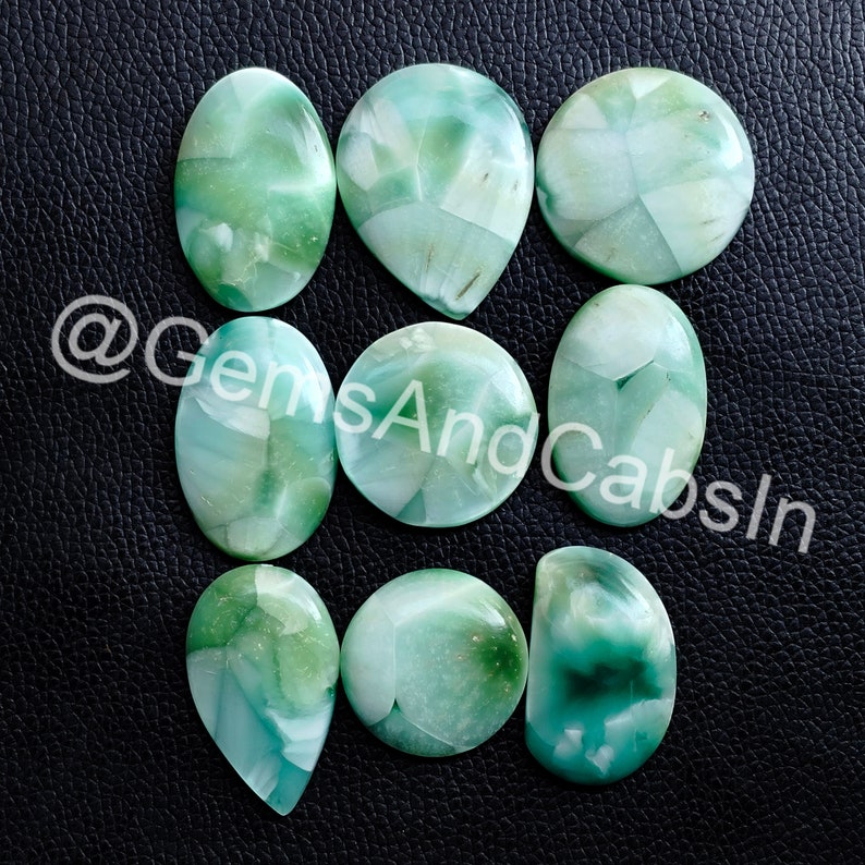 May include: Nine green and white cabochon gemstones with a swirling pattern. The gemstones are arranged in a grid pattern on a black surface.