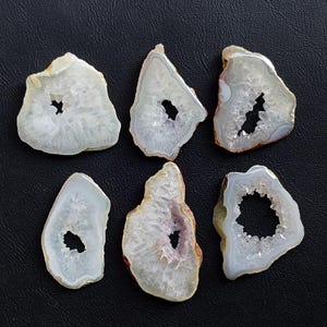 May include: Six polished agate slices with a geode centre are displayed on a black surface. The slices vary in shape and size, showcasing a range of colours from white to light grey, with some featuring hints of purple and brown.
