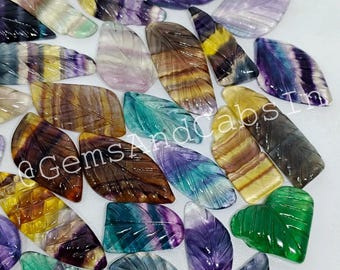 May include: A collection of carved gemstone leaves in various colors, including green, purple, yellow, and brown. The leaves are intricately detailed with veining and are arranged on a white surface. The text "@GemsAndCabsIn" is visible.