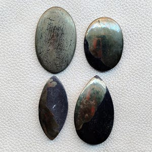 May include: Four polished gemstones in oval and teardrop shapes. The top two are metallic gray, while the bottom two are dark blue with iridescent patches. The stones are arranged on a white surface.