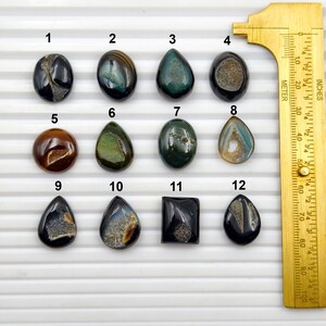 May include: A collection of twelve polished gemstones in various shapes and colors, including oval, teardrop, and square. The gemstones are displayed next to a ruler marked in inches and centimeters, with the longest gemstone measuring approximately 1.5 inches.