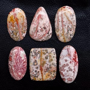 May include: Six polished gemstones in various shapes: oval, teardrop, and rectangular. The stones display a mix of colors, including cream, red, brown, and gray, with unique patterns and textures. The gemstones are arranged on a dark surface.
