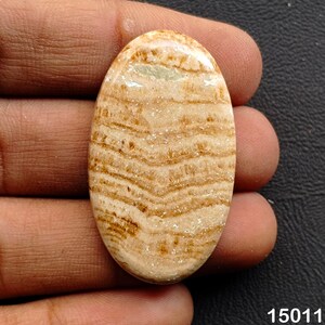 Puede incluir: Una gema ovalada con un patr&oacute;n en capas de tonos beige claro y marr&oacute;n. La superficie pulida revela estr&iacute;as naturales, creando una textura visual &uacute;nica. La gema se sostiene en una mano, mostrando su tama&ntilde;o y forma.