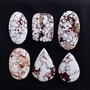 May include: Six white and brown gemstone cabochons in various shapes: oval, rectangular, and teardrop. The stones have a marbled appearance with brown veining and speckling against a white background. The cabochons are polished and smooth.