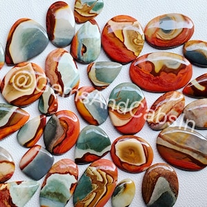 May include: A collection of 20 colourful, polished, and smooth, natural gemstone cabochons. The cabochons are a mix of red, orange, brown, green, and grey colours. The cabochons are all different shapes and sizes.