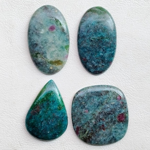 May include: Four polished gemstones in oval, teardrop, and square shapes. The stones have a mix of teal, green, and gray colors with red inclusions. The gemstones are likely for jewelry making or collecting.