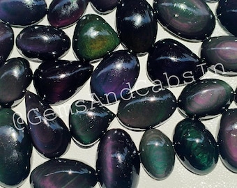 May include: A selection of polished obsidian gemstones in diverse shapes. The stones exhibit a spectrum of colours, including deep purples, greens, and blacks, with a glossy, reflective surface. The text "@GemsAndCabsIn" is visible.