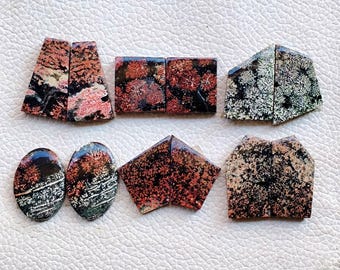 May include: Assortment of polished gemstones in various shapes, including trapezoids, squares, and ovals. The stones feature a mix of colors, including black, red, pink, and white, with intricate patterns. The gemstones are displayed on a white surface.
