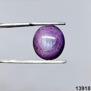 May include: A round, polished, purple gemstone held by tweezers. The gem has a star-like pattern on its surface. The gem is a deep purple colour with lighter purple and white highlights. The background is a light grey.