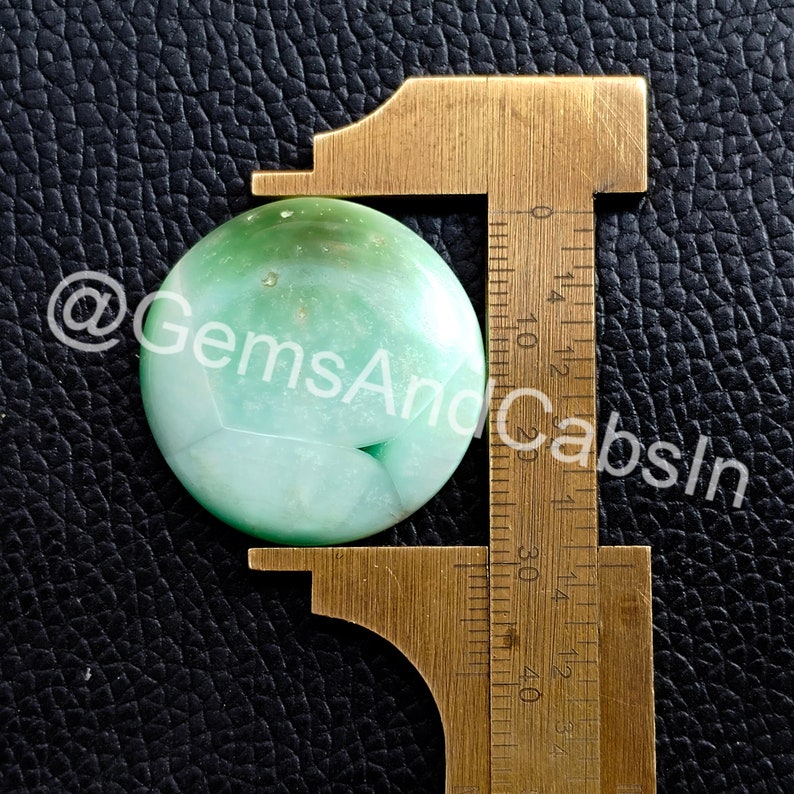 May include: A green and white agate cabochon, measuring approximately 2.5 cm in diameter, is displayed on a brass caliper.