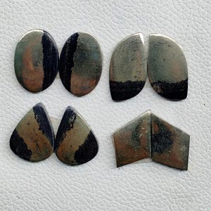 May include: Six polished gemstones in different shapes are displayed. There are two ovals, two half-moons, two teardrops, and two trapezoids. The stones have a mix of grey, brown, and black colours, with a striped or banded pattern. The gemstones are presented on a white surface.