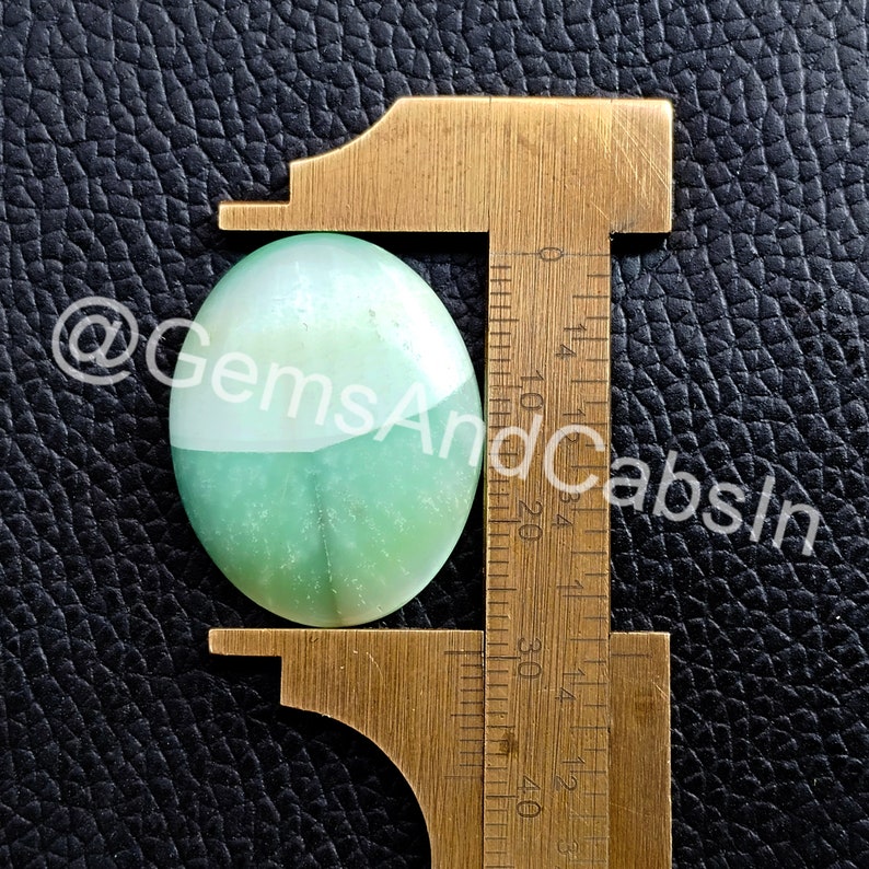 May include: A green and white oval cabochon gemstone measuring approximately 2.5 cm in length, displayed on a ruler.