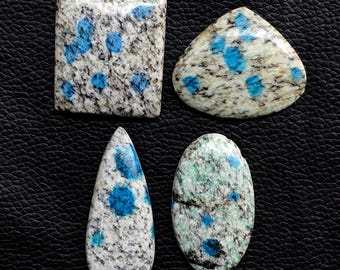 May include: Four polished K2 stone cabochons in various shapes: a rectangle, a triangle, a teardrop, and an oval. The stones have a speckled white and gray base with vibrant blue spots. The stones are displayed on a black surface.