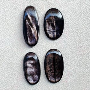 Puede incluir: Cuatro piedras preciosas de obsidiana negra pulida con patrones iridiscentes. Dos son de forma ovalada y dos son de forma ovalada alargada. Las piedras tienen un acabado brillante y se muestran sobre una superficie blanca.