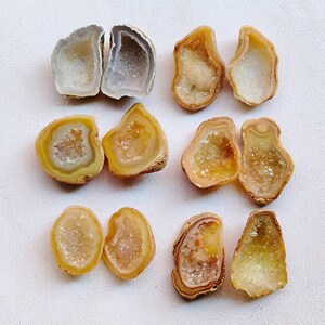 Puede incluir: Una colección de ocho mitades de geodas en varios colores, incluyendo blanco, gris, amarillo y marrón. Cada geoda está abierta, revelando un interior de cristal brillante. Las geodas están dispuestas sobre una superficie blanca.