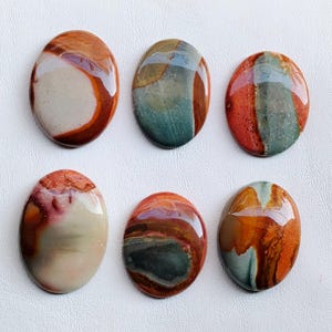 May include: Six polished, oval gemstones in shades of brown, red, cream, and teal. Each stone has a unique pattern and glossy finish. The gemstones are arranged on a white surface.