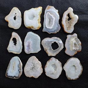 May include: A collection of twelve polished agate slices, each with a unique shape and internal crystal formation. The slices display a range of colors, including white, gray, and hints of purple, with a contrasting brown outer edge. The geode slices are arranged on a black surface.