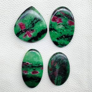 May include: Four polished ruby zoisite gemstones in various shapes. The stones feature a vibrant green base color with black veining and patches of deep red ruby. One stone is teardrop-shaped, while the others are oval.