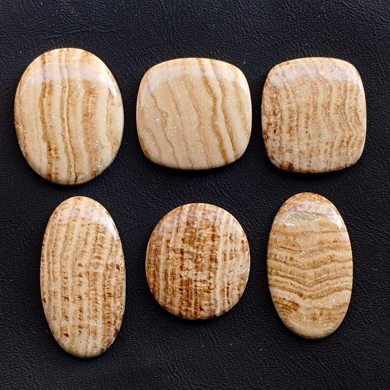 Puede incluir: Seis piedras pulidas de color beige con vetas marrones dispuestas sobre una superficie negra. Las piedras tienen varias formas: ovalada, redonda y cuadrada con esquinas redondeadas. El veteado crea un patr&oacute;n natural y org&aacute;nico.