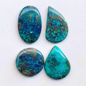 May include: Four polished gemstones in various shapes: oval, teardrop, half-moon, and round. The stones feature a mix of blue, teal, and brown colors with unique veining patterns. The gemstones are displayed on a white surface.