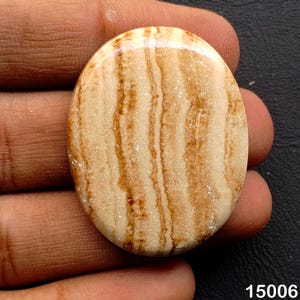 Puede incluir: Gema ovalada pulida con base crema y bandas marrones onduladas. La piedra se sostiene en una mano. El n&uacute;mero "15006" es visible en la esquina inferior derecha. El patr&oacute;n de la gema se asemeja a la veta de la madera.