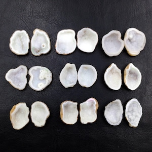 May include: A collection of eighteen small, irregular, white geode slices arranged in three rows of three pairs. Each piece features a hollow centre filled with sparkling crystalline formations and a smooth, rounded outer edge in shades of cream.