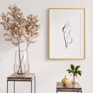 May include: A gold framed print with a black line drawing of a person. The print is hanging on a white wall above a brown wooden table with a clear glass vase filled with dried branches. A small gold vase with a flower arrangement is on the table.