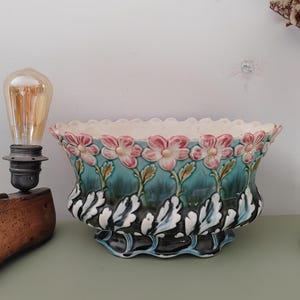Puede incluir: Una jardinera de cerámica con borde festoneado, con flores rosas y hojas verdes sobre un fondo turquesa. La base tiene un diseño de hojas en blanco y negro. Decorativa y vintage.