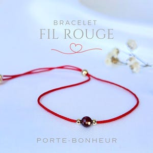 Könnte beinhalten: Rotes Armband mit einer kleinen roten Edelsteinperle und goldenen Perlen. Das Armband ist mit einem Knoten gebunden und hat an einem Ende einen Quasten. Der Text "BRACELET FIL ROUGE PORTE-BONHEUR" ist auf weißem Hintergrund geschrieben.