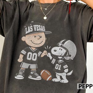 Puede incluir: Una camiseta gris carbón de manga corta con un gráfico de Charlie Brown con una gorra de Las Vegas y Snoopy con un casco de fútbol americano. Ambos personajes llevan camisetas con los números 00 y 01, chocando los puños junto a un balón marrón.