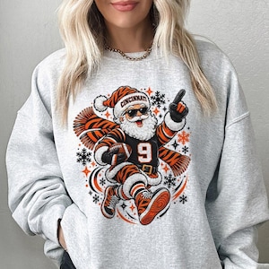 May include: A light grey crewneck sweatshirt featuring a graphic of Santa Claus wearing sunglasses, a tiger-striped scarf, and a jersey with the number 9. Santa holds a football and points upward, surrounded by orange stars and black snowflakes.