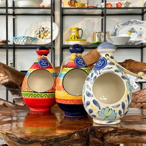 May include: Three ceramic salt cellars with a unique teardrop shape and a hole in the side. The cellars are decorated with colorful patterns and designs, including blue, red, yellow, green, and white. Each cellar has a rope handle.